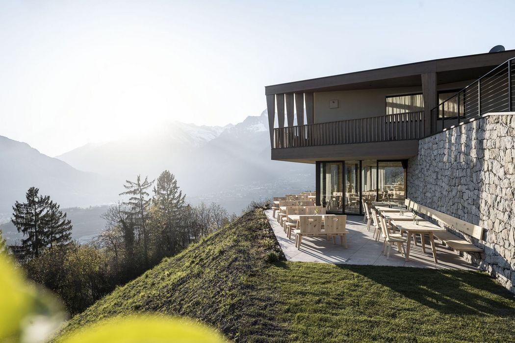 Moderner Außenbereich eines Hauses mit Holzmöbeln auf einer Terrasse, umgeben von Hügeln und Bergen im Sonnenlicht. Moderner Außenbereich eines Hauses mit Holzmöbeln auf einer Terrasse, umgeben von Hügeln und Bergen im Sonnenlicht.