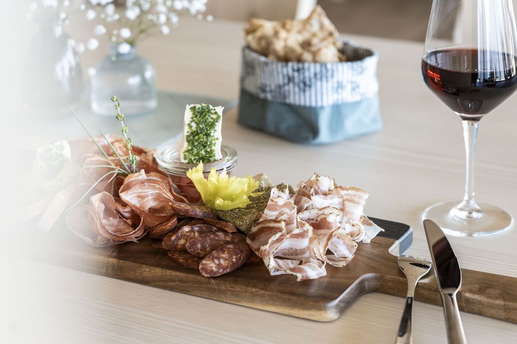 Holzbrett mit verschiedenen Wurst- und Käsesorten, Kräutern, Brot im Hintergrund und Glas Rotwein auf einem Tisch Holzbrett mit verschiedenen Wurst- und Käsesorten, Kräutern, Brot im Hintergrund und Glas Rotwein auf einem Tisch