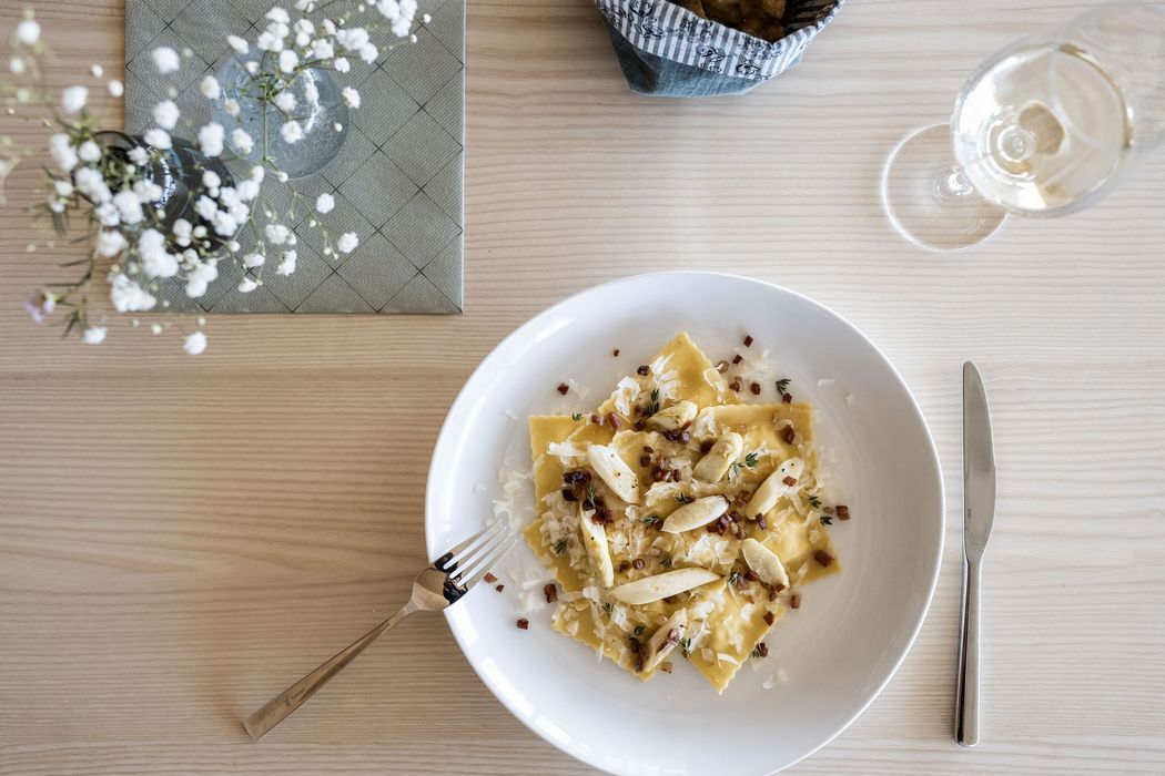 Teller mit Ravioli, Spargelstücken, Käse und Speck auf einem Holztisch mit Besteck, Glas und Blumen in der Vase Teller mit Ravioli, Spargelstücken, Käse und Speck auf einem Holztisch mit Besteck, Glas und Blumen in der Vase