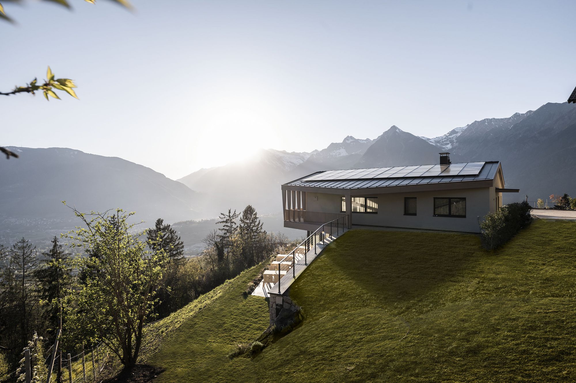 Modernes Haus mit Solarpanels auf dem Dach, umgeben von grüner Wiese und Bergen im Sonnenuntergang Modernes Haus mit Solarpanels auf dem Dach, umgeben von grüner Wiese und Bergen im Sonnenuntergang
