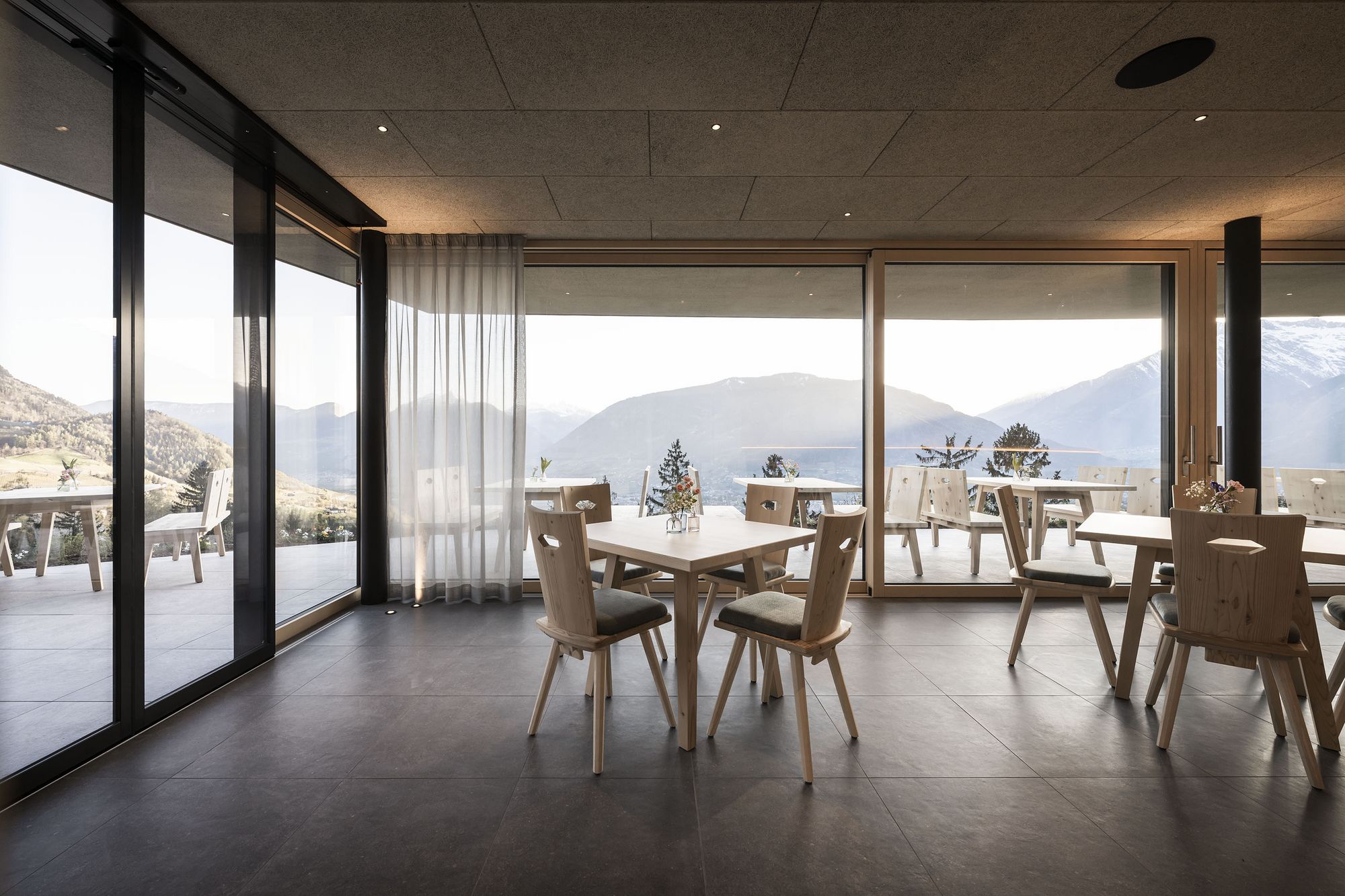Modernes Restaurant mit Holzstühlen und Tischen, großer Glasfront mit Bergpanorama Modernes Restaurant mit Holzstühlen und Tischen, großer Glasfront mit Bergpanorama