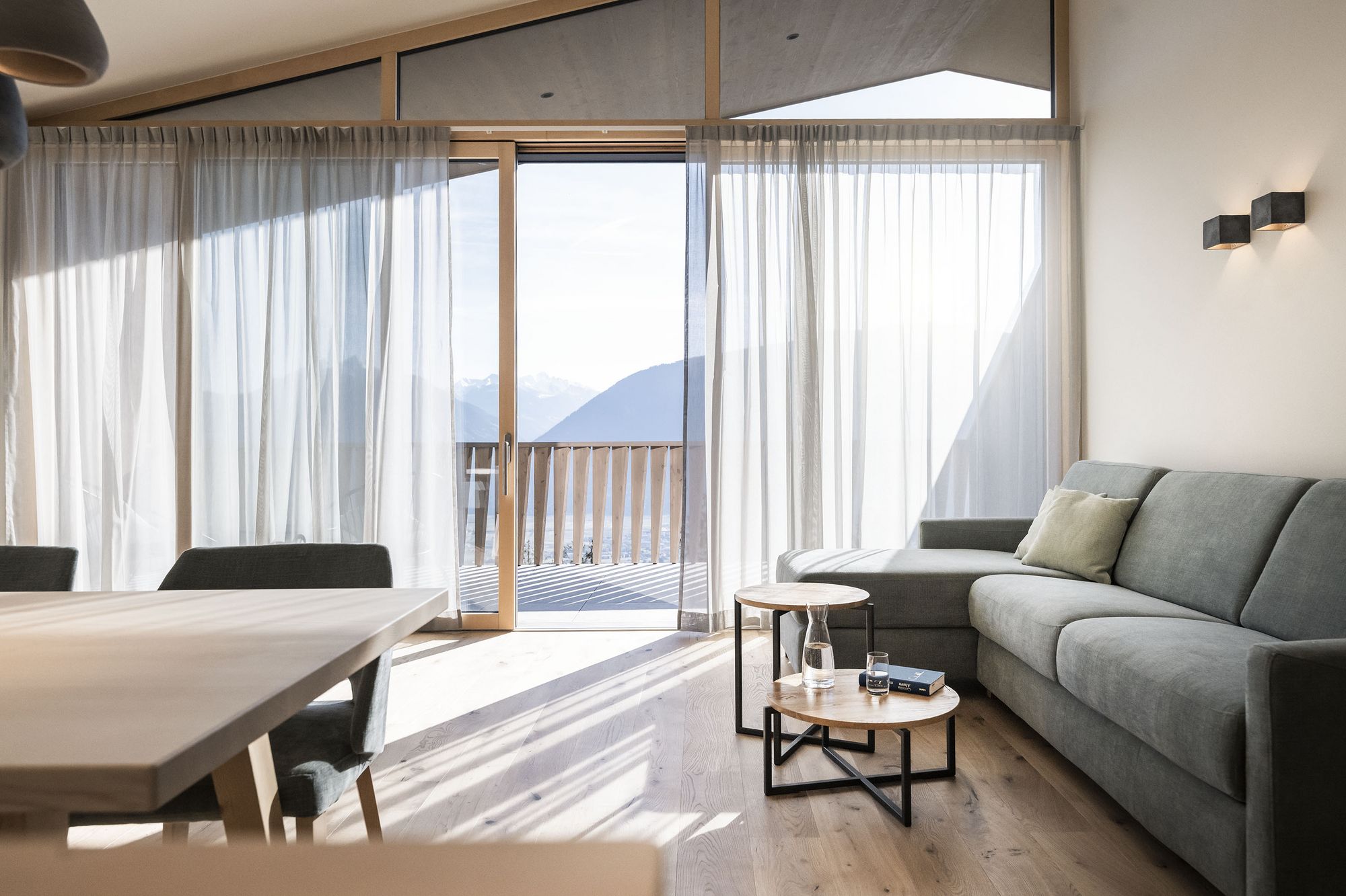 Heller, moderner Wohnraum mit großem Sofa, Holztisch, durchscheinenden Vorhängen und Blick auf Berglandschaft durch große Fenster. Heller, moderner Wohnraum mit großem Sofa, Holztisch, durchscheinenden Vorhängen und Blick auf Berglandschaft durch große Fenster.