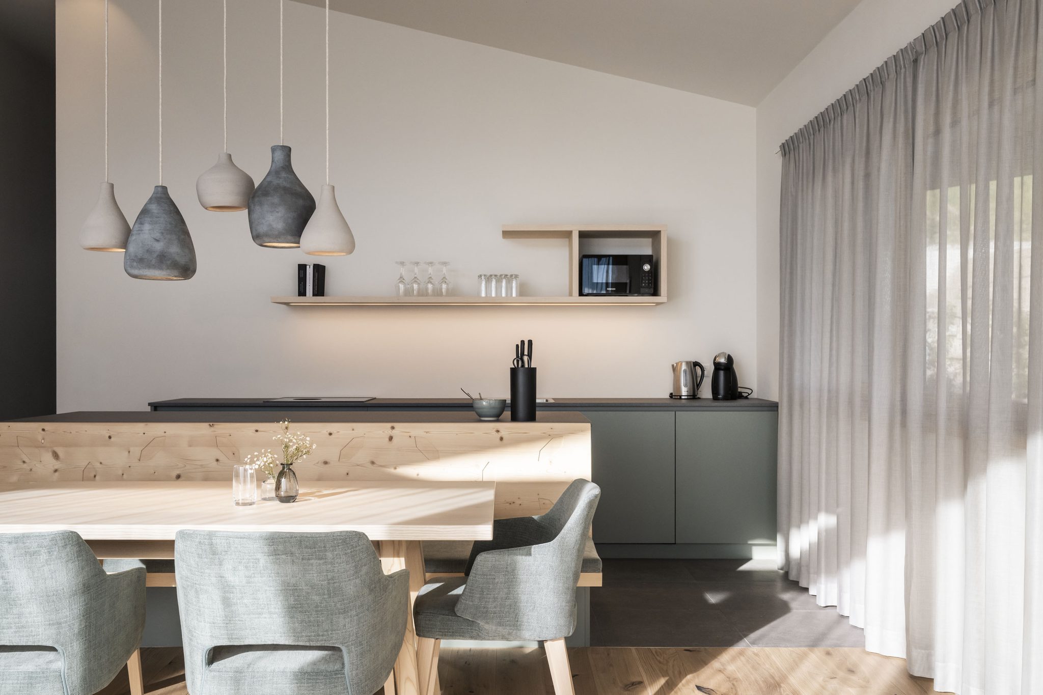 Moderne minimalistische Küche mit Holztisch, grauen Stühlen, hängenden Keramiklampen und Küchenzeile mit Geräten, heller Raum mit Vorhängen Moderne minimalistische Küche mit Holztisch, grauen Stühlen, hängenden Keramiklampen und Küchenzeile mit Geräten, heller Raum mit Vorhängen