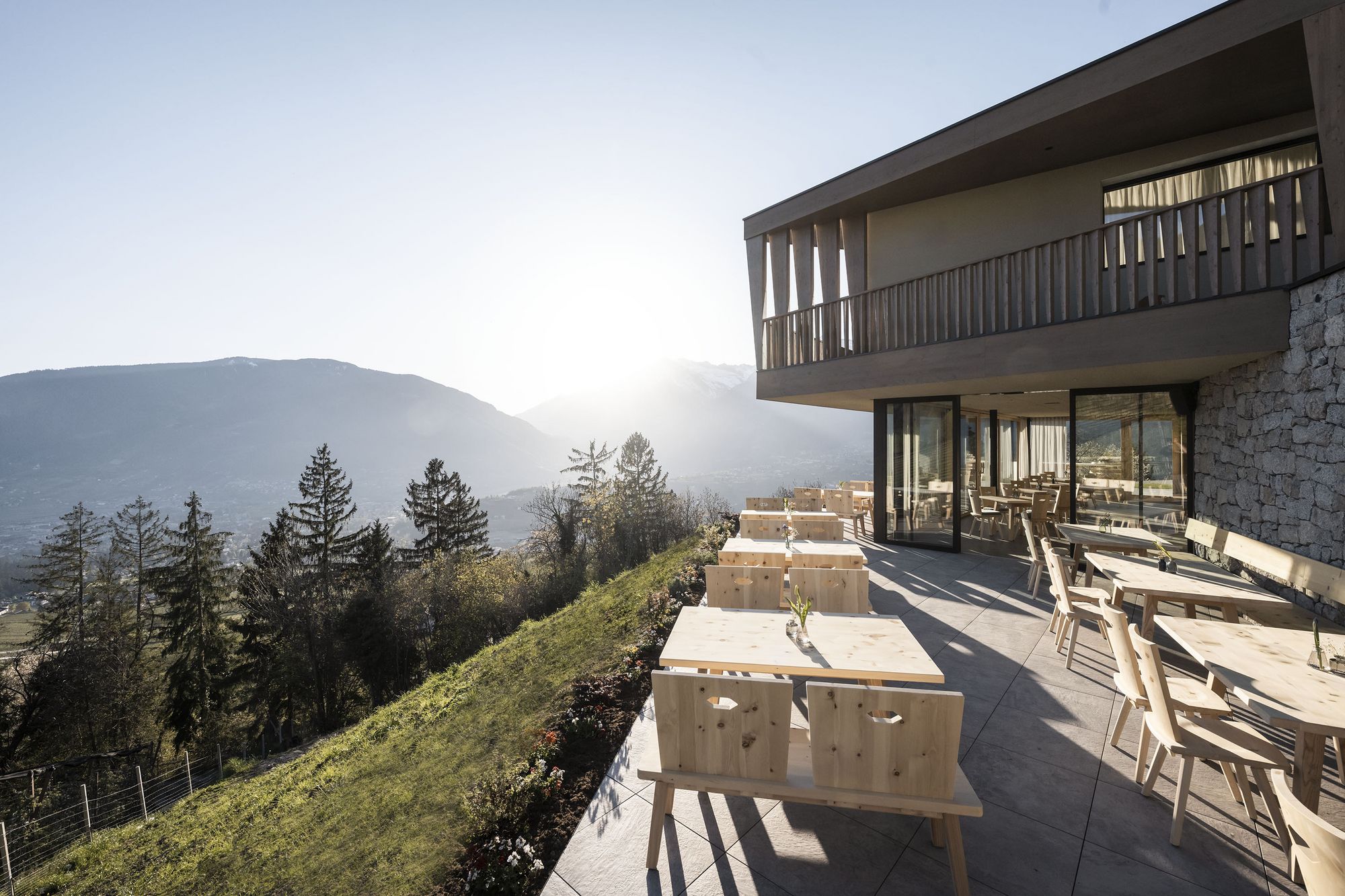 Terrasse mit Holztischen und Stühlen neben modernem Gebäude mit Blick auf Berge und Sonnenuntergang Terrasse mit Holztischen und Stühlen neben modernem Gebäude mit Blick auf Berge und Sonnenuntergang